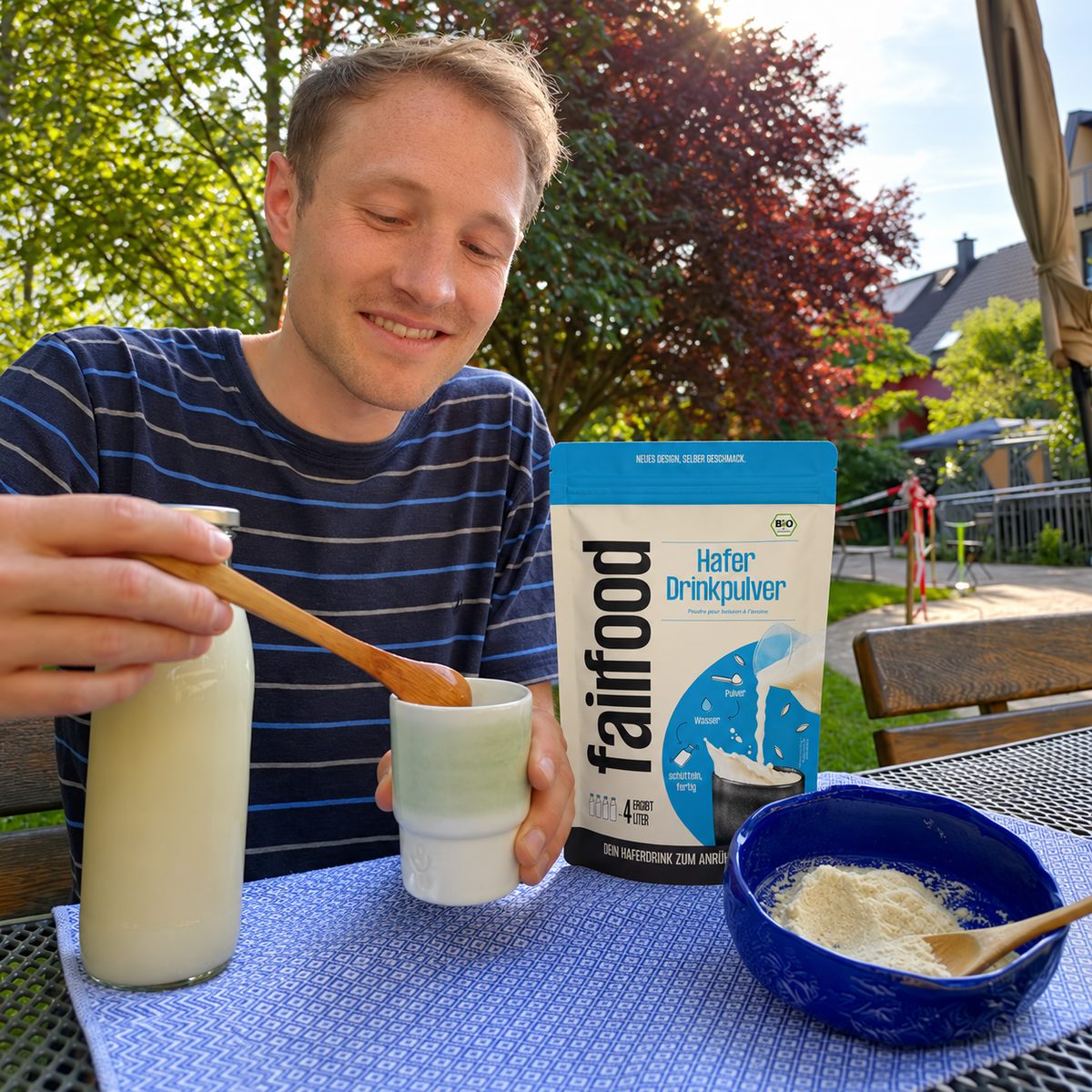 Benedikt rührt Haferdrink in einem Keramikbecher an; daneben stehen Haferdrinkpulver, eine Glasflasche und eine Schale mit Pulver