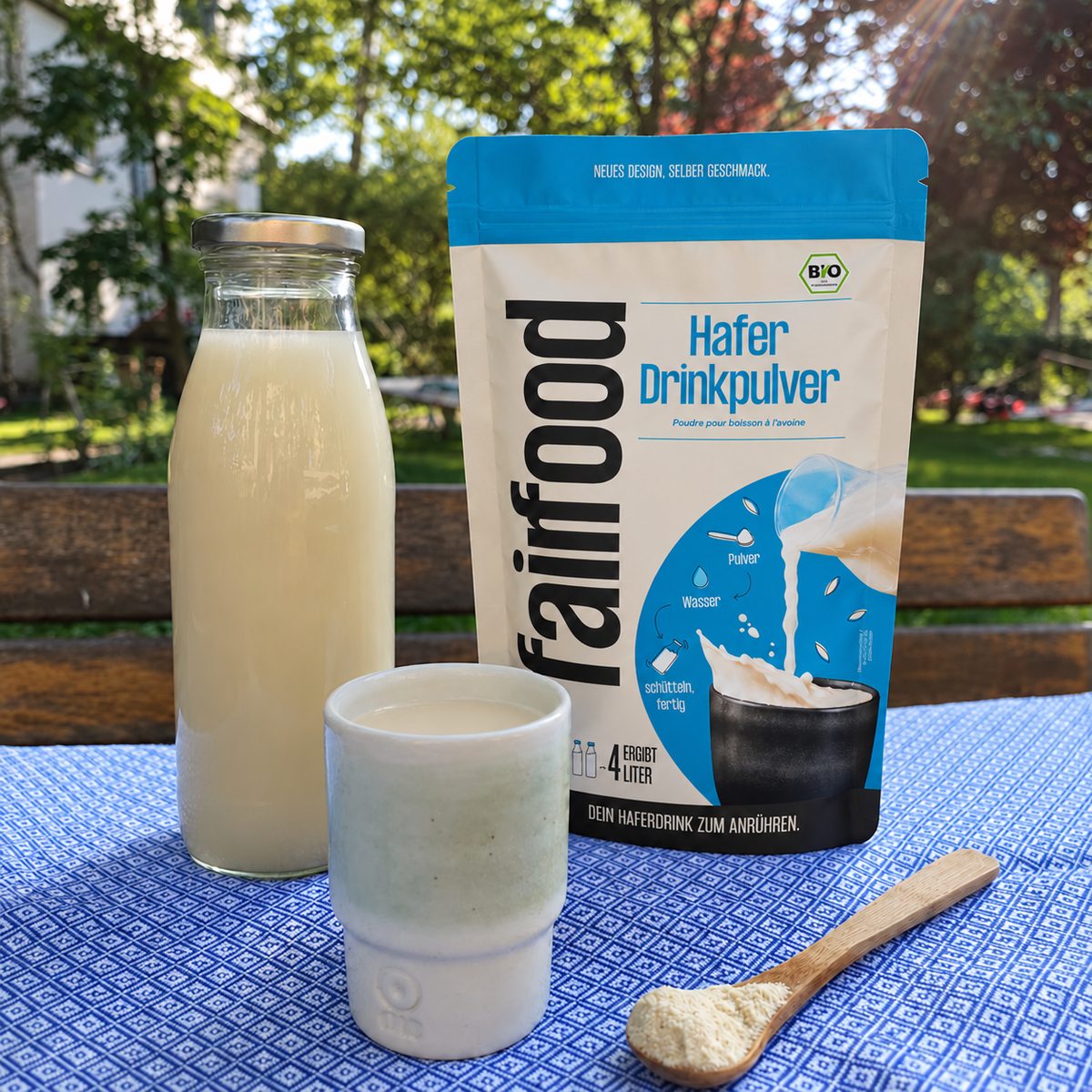 Becher mit cremig hellem Haferdrink vor einem Beutel Haferdrinkpulver, daneben eine Glasflasche und ein Holzlöffel mit Pulver.